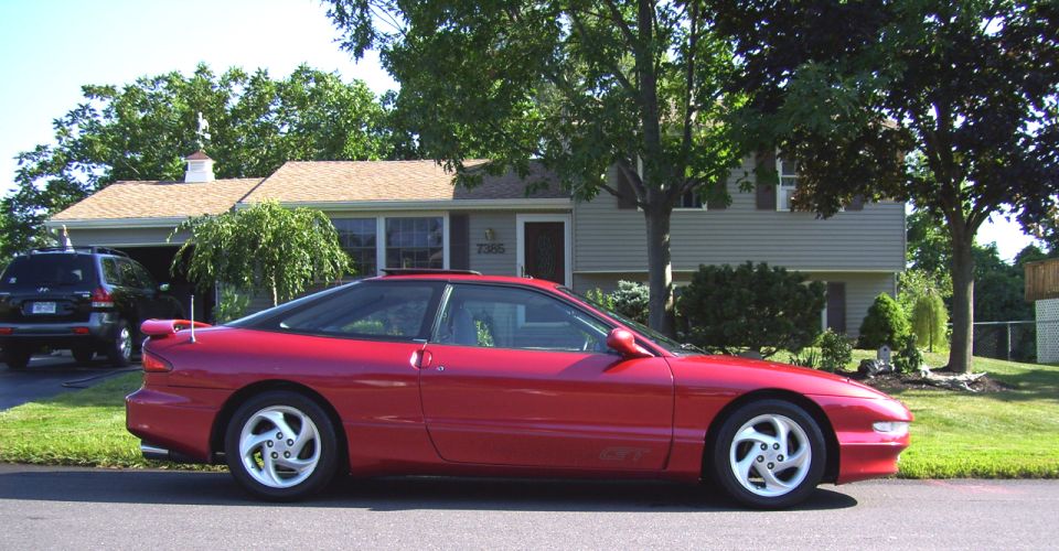 Red 1995 Ford Probe GT parked on street, black SUV in driveway behind it