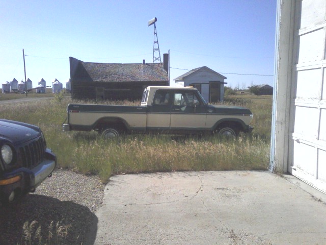 Green and tan Ford pickup truck parked in tall grass beside rural buildings, side view