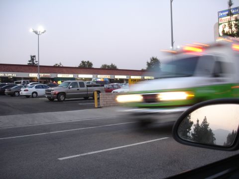 1993 Ford Taurus - An ambulance with flashing lights speeds by a parking lot in front of a strip mall.