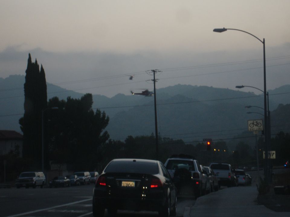 Two helicopters fly over a road with multiple cars in traffic at dusk