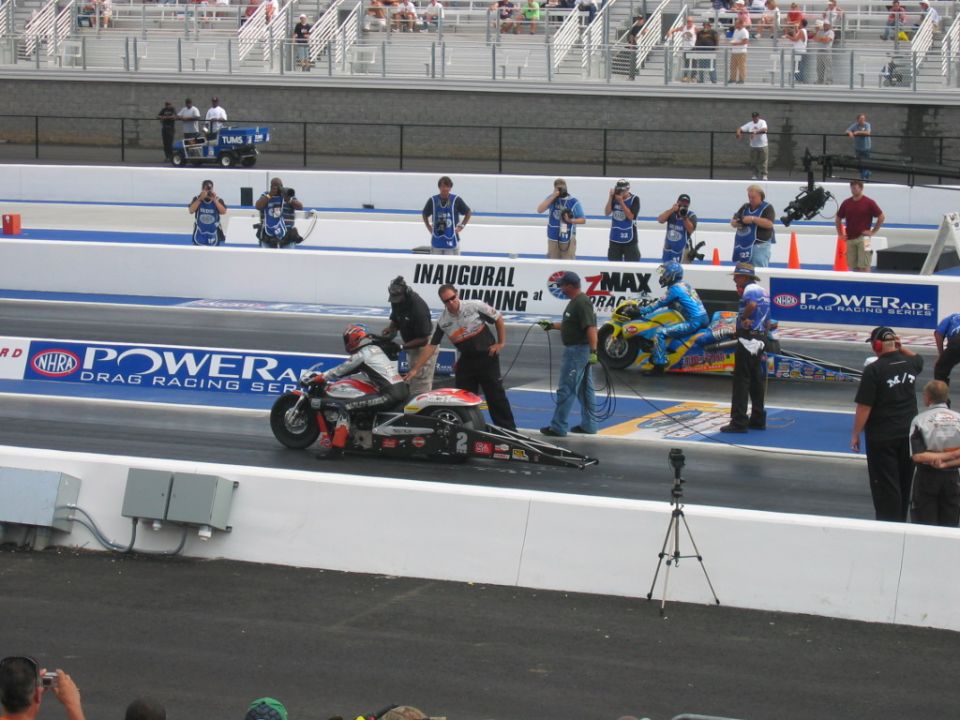 Drag racing motorcycles on a track with crew members and spectators in the background