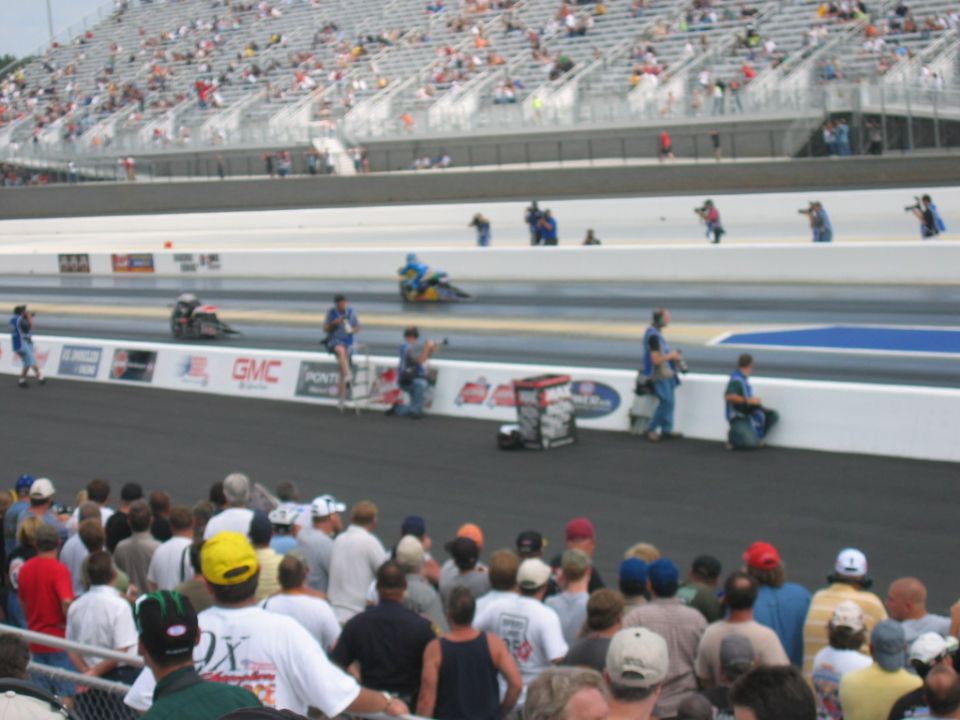 Spectators watch drag racers on a track with grandstands in the background