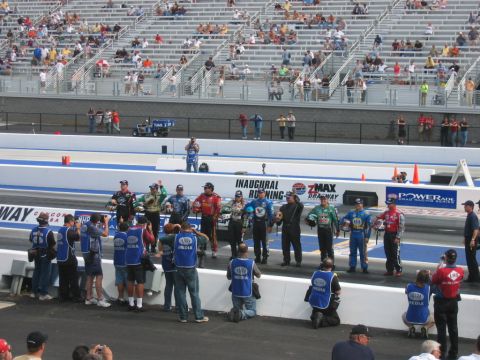 1992 Ford Mustang - A group of drag racers and media members are gathered on a drag strip with spectators in the stands.