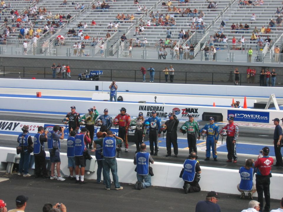 Drag racers in uniform stand on a raised platform with media personnel below, grandstands in background