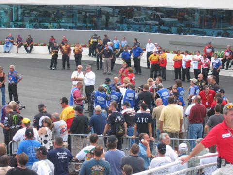 1992 Ford Mustang - A large group of people, some in official vests, are gathered on the asphalt at a race track, with a building and vehicles visible in the b…