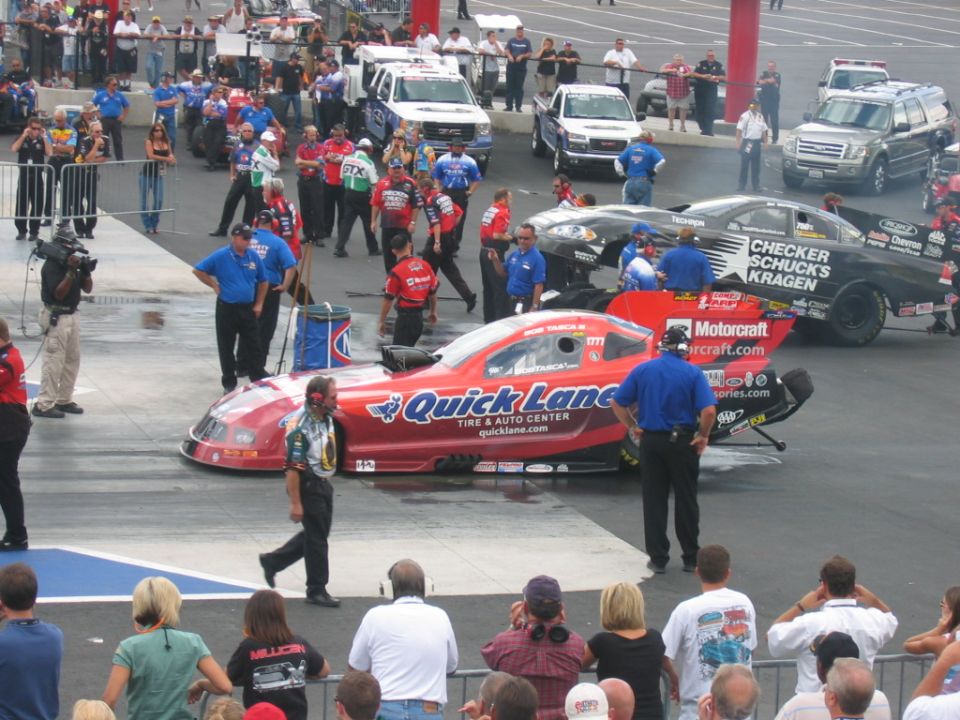 Red and black drag racing cars on a track with crews and spectators
