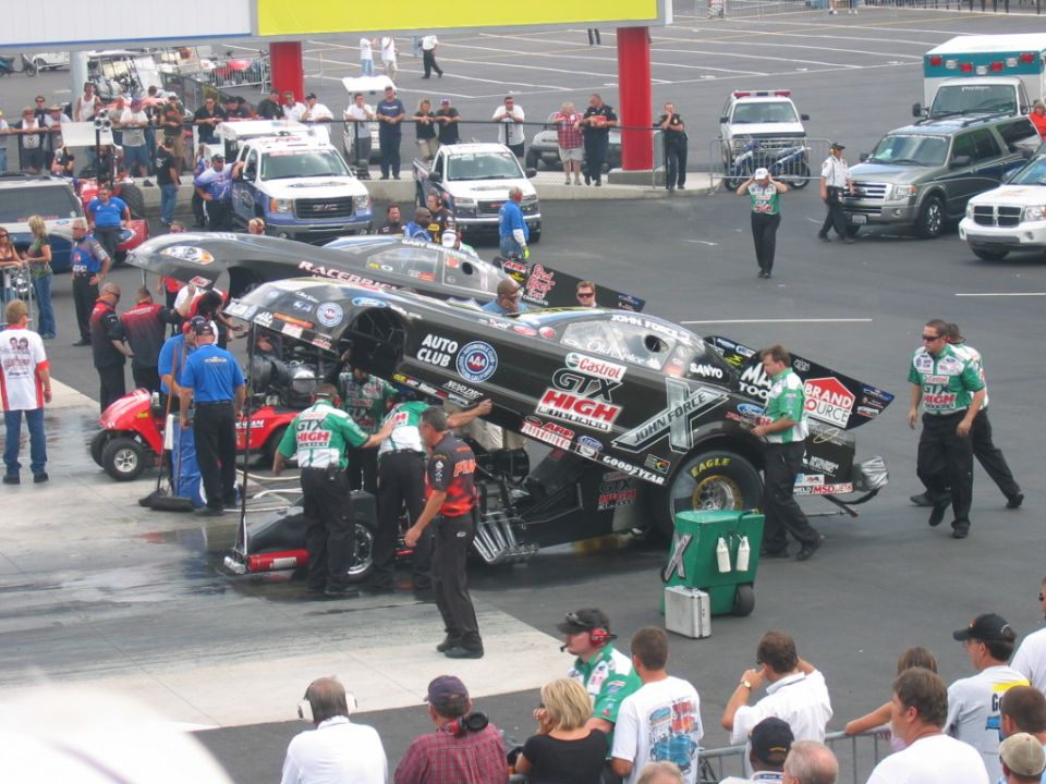Two drag racing cars with crew members working on them, one black with "GTX HIGH" livery, the other partially visible