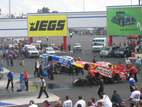 1992 Ford Mustang - Crew members work on drag racing cars at a track, with sponsor banners overhead.