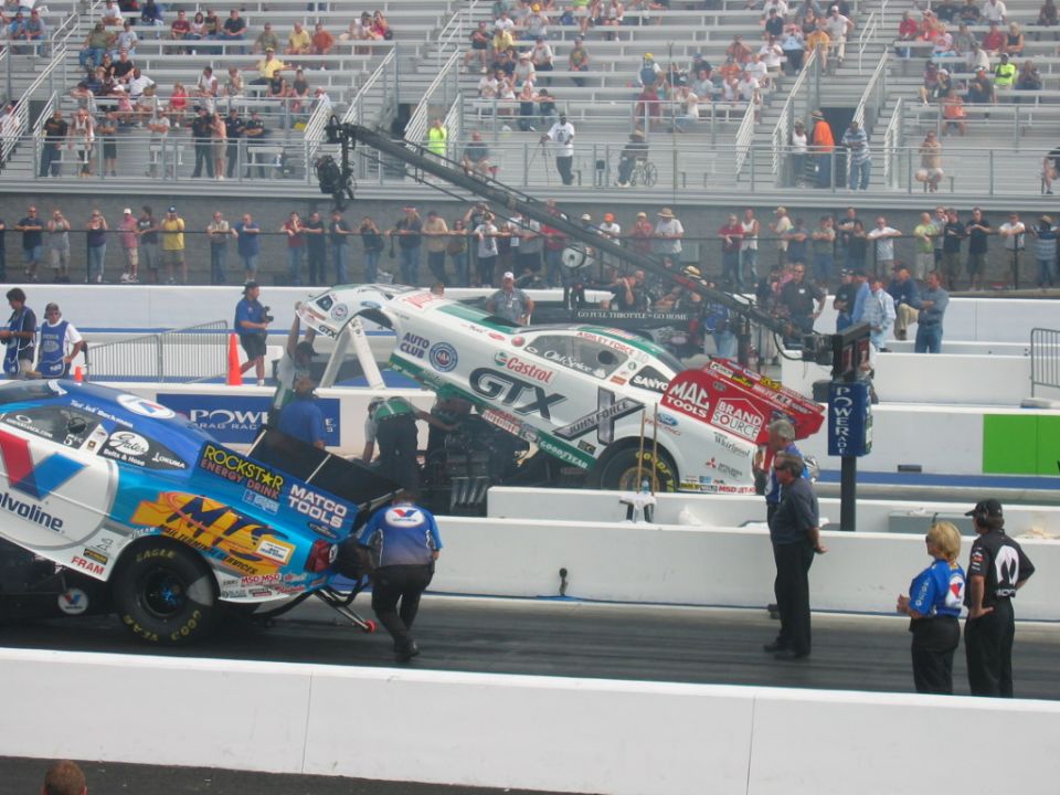two drag cars on track, one white with green trim, other blue with white trim, people around