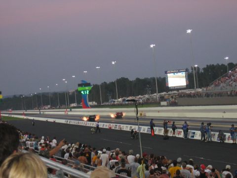 1992 Ford Mustang - Drag cars race down the track at dusk, with flames erupting from one vehicle and a crowd watching from the stands.