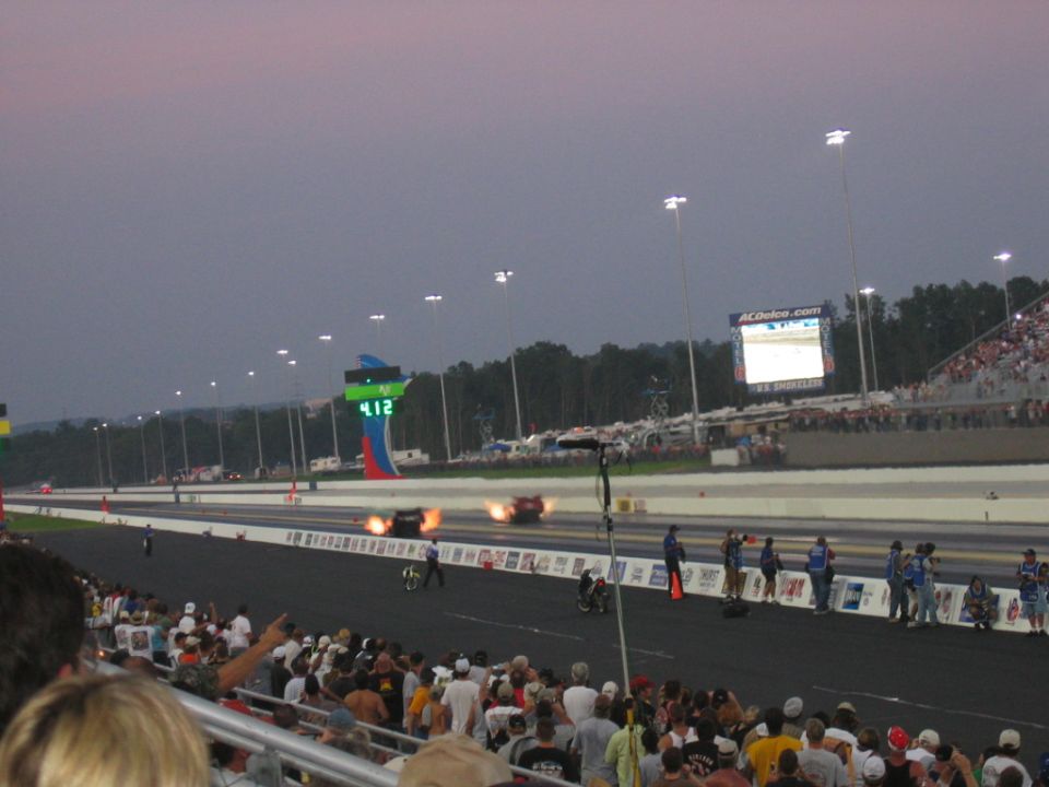 Drag racing cars on a track with flames, spectators in the foreground, and a large scoreboard in the background