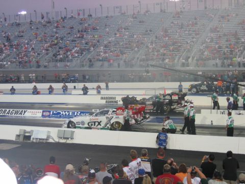 1992 Ford Mustang - Two drag racing cars are positioned at the starting line, with a crowd of spectators filling the grandstands in the background.