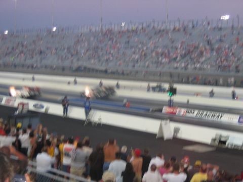 1992 Ford Mustang - Two drag cars compete on a track with a large crowd watching from the stands.