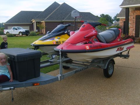 1994 Ford Bronco - Two jet skis are parked on a trailer in front of a suburban home, brick house.