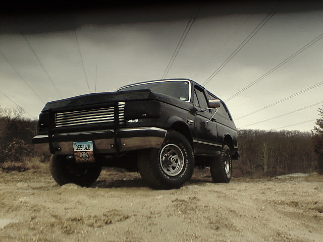 Black Ford Bronco 4x4 with brush guard and white-letter tires on sandy ground, shot from a low front angle