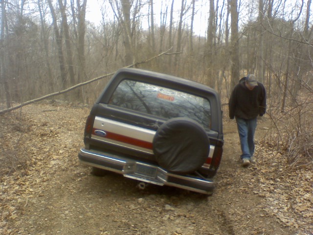 Rear of dark Ford Bronco with tailgate spare tire on wooded dirt trail, man walking beside it