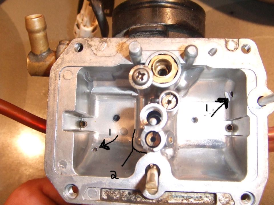 Close-up of a disassembled carburetor with arrows pointing to internal components. A hand is visible holding the part