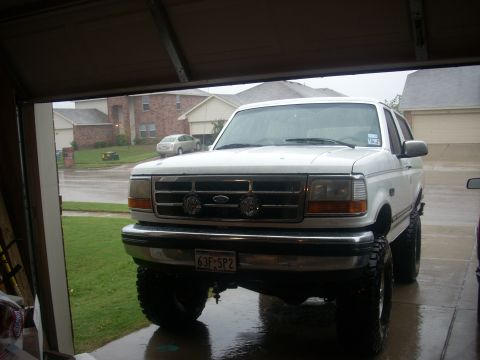 1992 Ford Bronco - Front view of a lifted white full-size Ford Bronco parked on a wet driveway in the rain, shot from inside an open garage. Two covered KC-st…