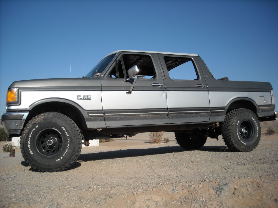 Silver lifted Ford crew-cab pickup, side view, topless rear on gravel