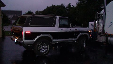 1979 Ford Bronco - A two-tone Ford Bronco is parked on a wet surface next to a large semi-truck.