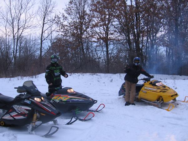 Three snowmobiles parked in snowy woods at dusk with two helmeted riders standing by