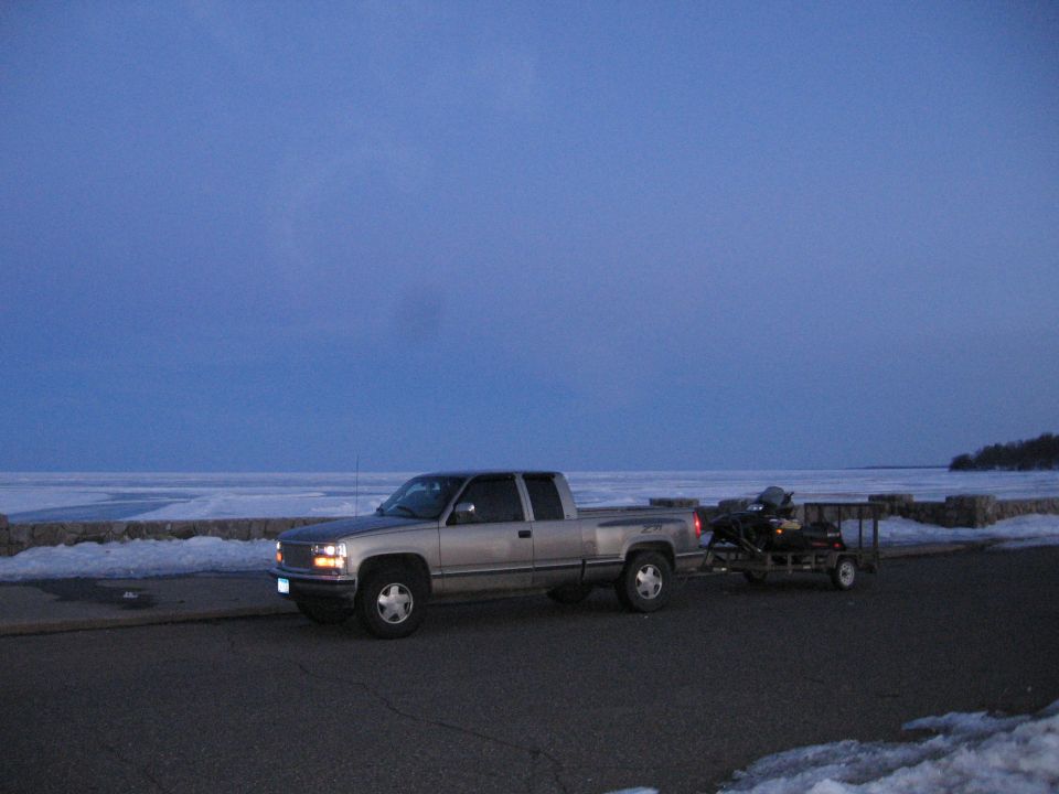 Tan pickup towing a snowmobile on a small trailer beside a frozen lake at dusk
