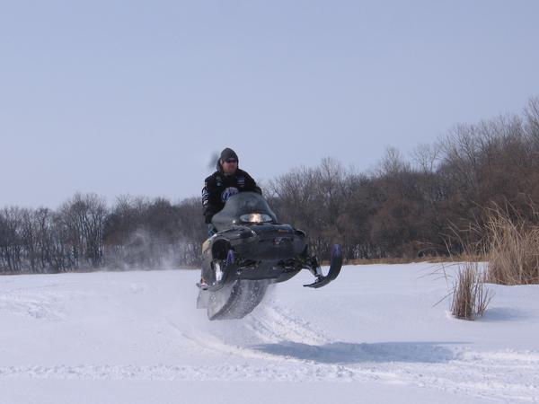 Rider jumping a black snowmobile off a snow bank under clear blue winter sky