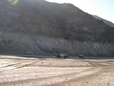 2007 Toyota FJ Cruiser - Two vehicles traverse a dry riverbed under a large, rocky hillside.