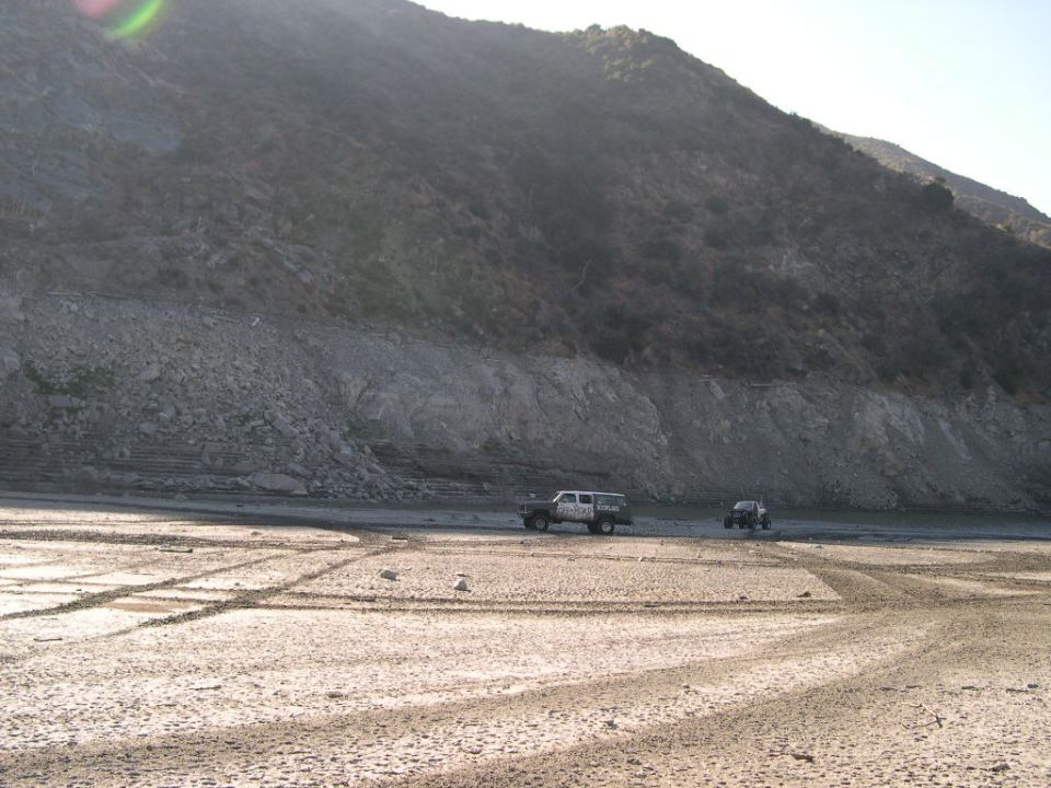 Two off-road vehicles driving on a dry, sandy riverbed with tire tracks, a large rocky hill in the background