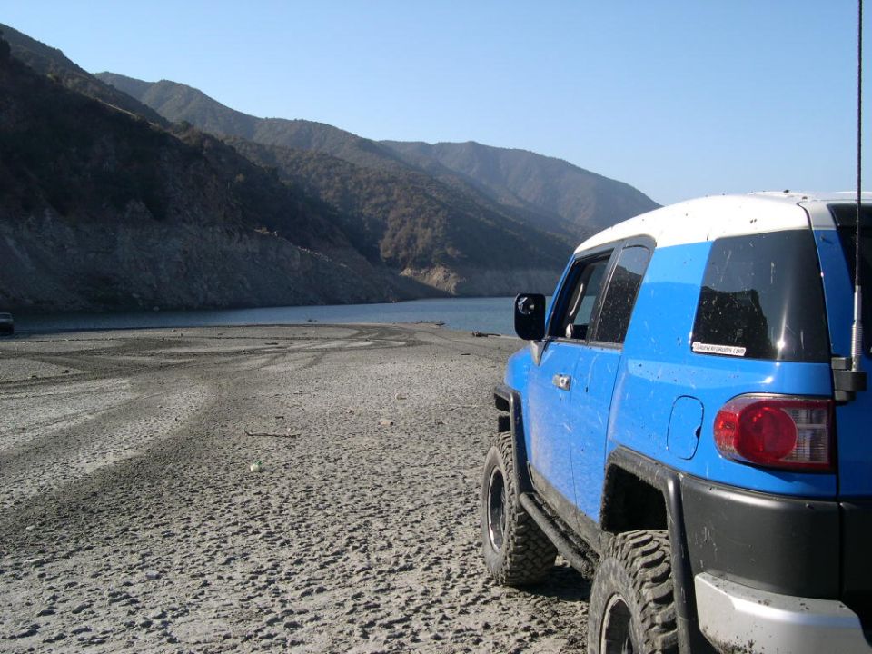 Blue 2007 Toyota FJ Cruiser with white roof parked on a dry, sandy shore next to a body of water