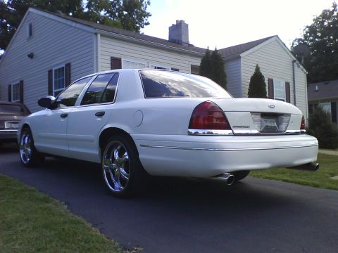 2003 Ford Crown Victoria - A white 2003 Ford Crown Victoria is parked in a driveway in front of a residential house. The car is shown from the rear passenger side qua…