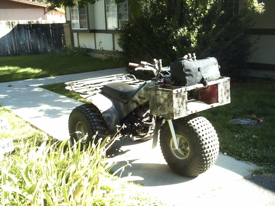 Camo-painted three-wheeler ATC with a front wire basket and duffel bag parked on a sidewalk
