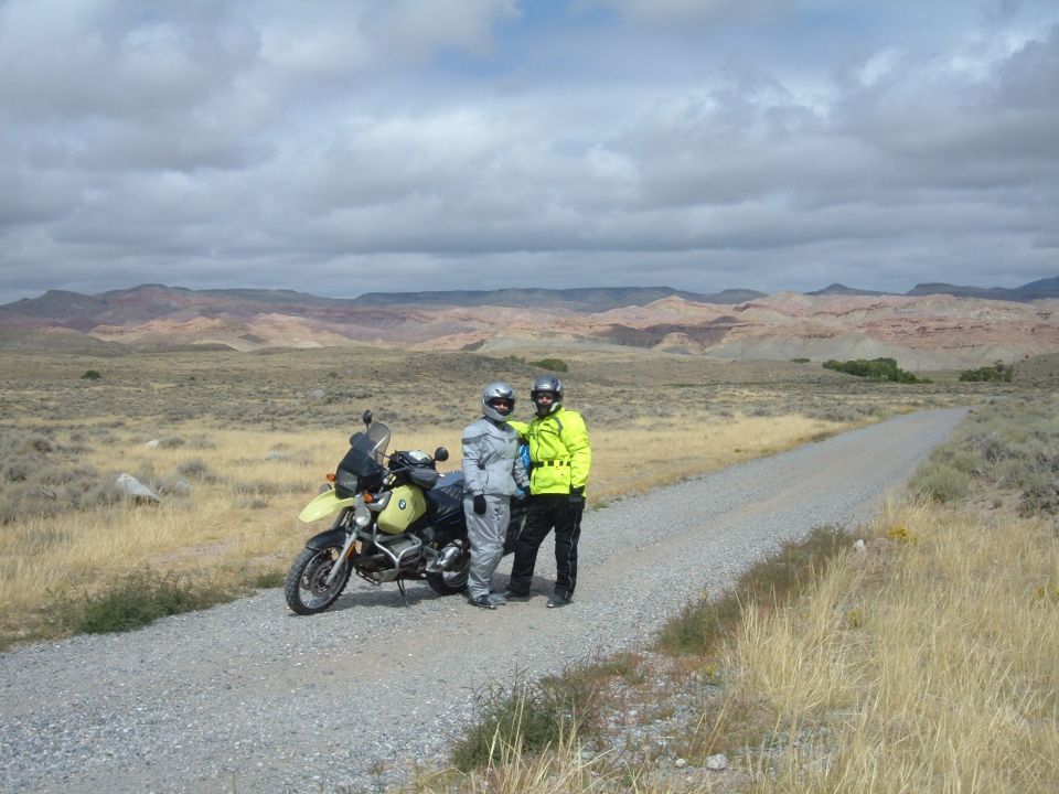 Two motorcyclists and a yellow BMW R1100GS on a gravel road in a desert landscape.