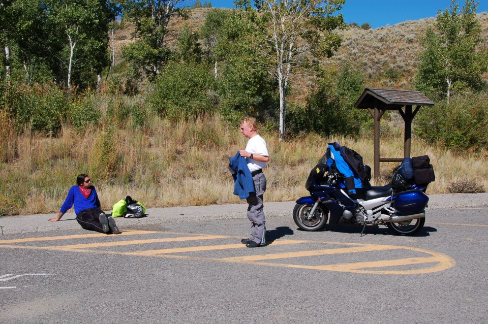 Blue touring motorcycle parked on road with two people nearby. One person sits on the ground, the other stands holding a jacket