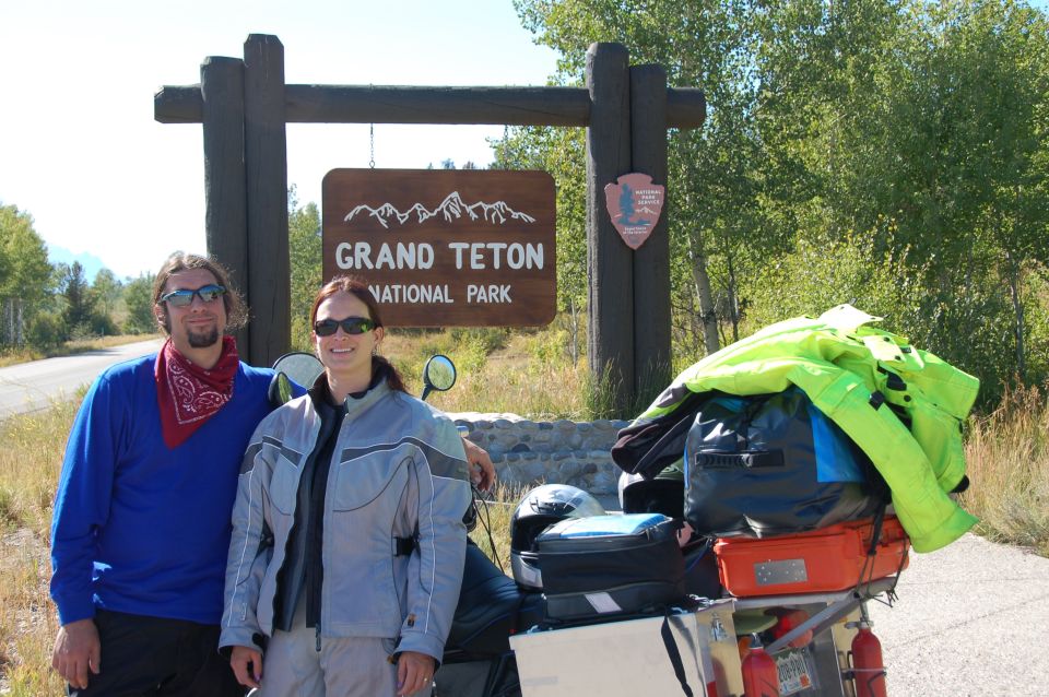 Man and woman at Grand Teton National Park sign with a loaded motorcycle.