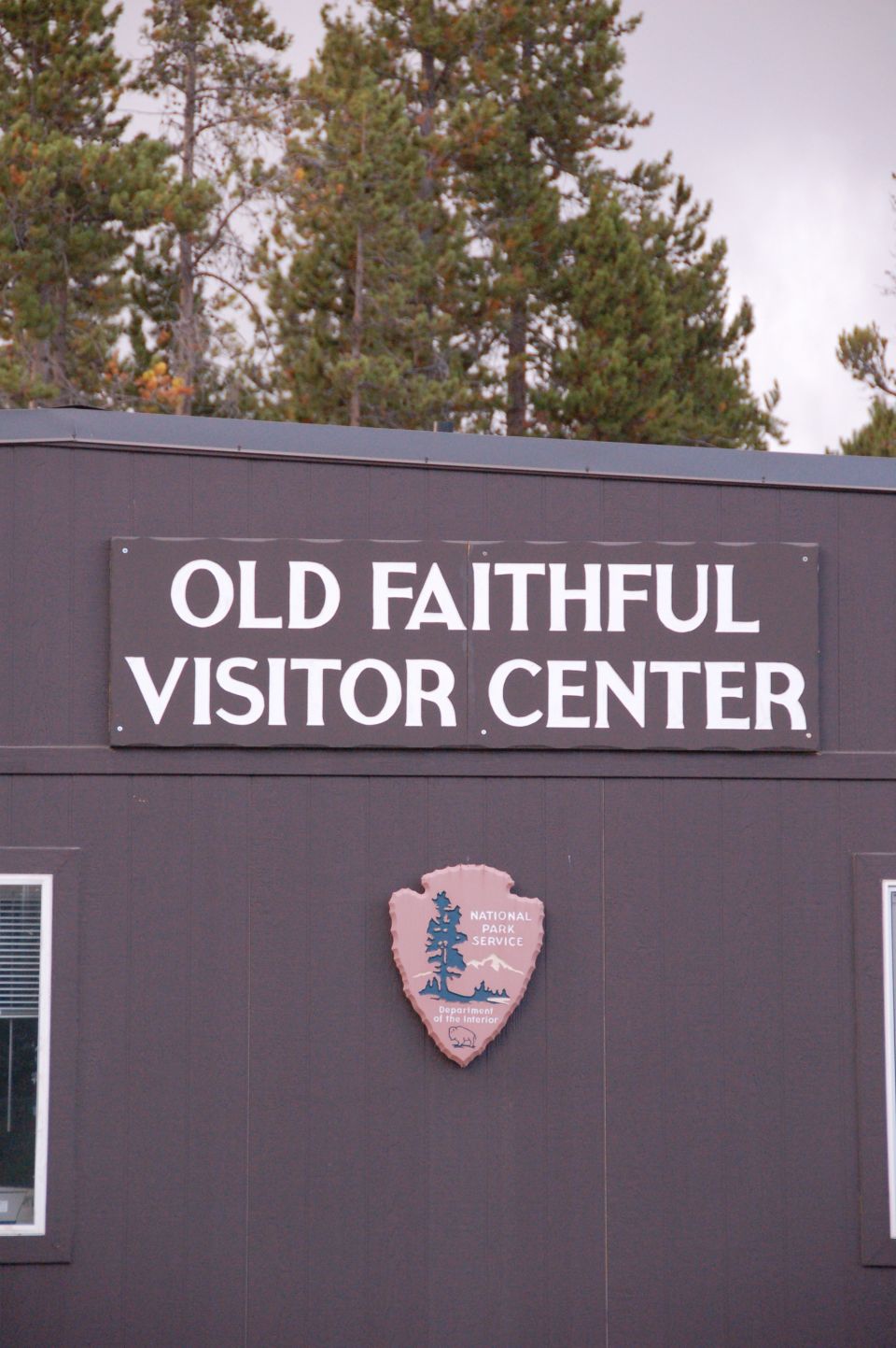 Sign for Old Faithful Visitor Center on a building with pine trees.