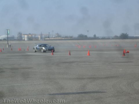1985 Ford Mustang - A silver-grey two-seat sports coupe runs an autocross course on a wide asphalt lot lined with orange traffic cones, tire smoke trailing beh…