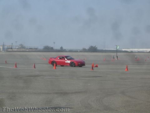 1985 Ford Mustang - A red Mazda RX-7 is driving on a paved track marked with orange cones. The car has the number "877" visible on its door.