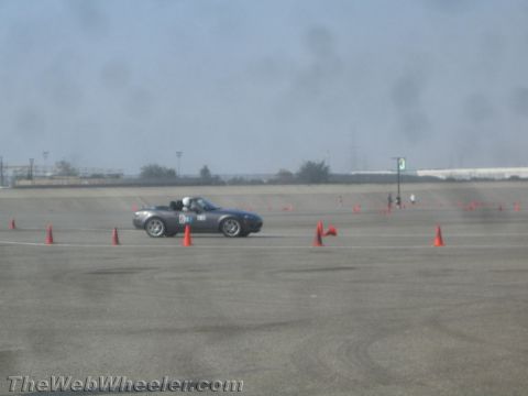 1985 Ford Mustang - A gray convertible sports car with a driver wearing a helmet navigates a course marked by orange cones on a paved surface. The car has a ra…