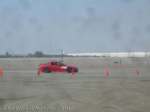 1985 Ford Mustang - A red sports car with the number "877" on its door is driving on a paved track, navigating around orange cones.