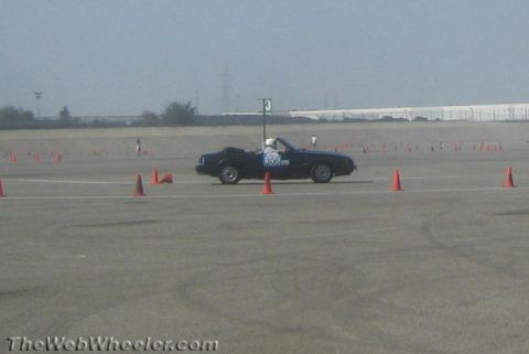 1985 Ford Mustang - A black convertible Mustang is driving through a cone course on a paved lot. The car has the number 306 displayed on its door.