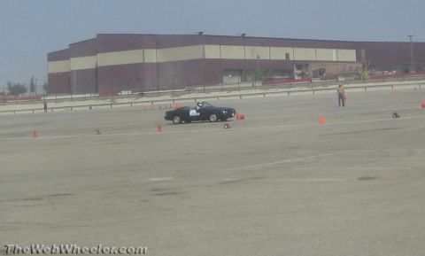 1985 Ford Mustang - A black convertible Mustang is shown on a paved lot, navigating a course marked by orange cones. A person in blue jeans and no shirt is vis…