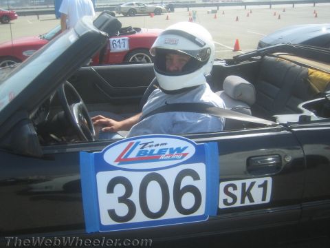 1985 Ford Mustang - A driver wearing a white helmet sits in the driver's seat of a black convertible Mustang, ready for a race. Visible on the car door are rac…