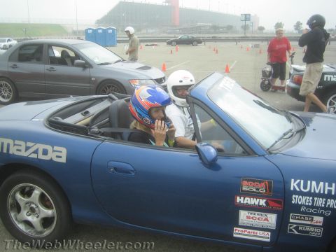 1985 Ford Mustang - A blue Mazda Miata convertible with two people wearing racing helmets is parked on a track. Various racing decals are visible on the car's…