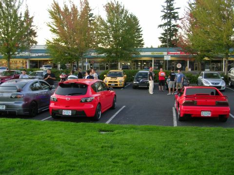 2007 Mazda 3 - A red 2007 Mazda 3 hatchback is parked in a lot with other vehicles and people.