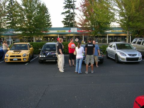 2007 Mazda 3 - A group of people stand around cars in a parking lot in front of a strip mall.