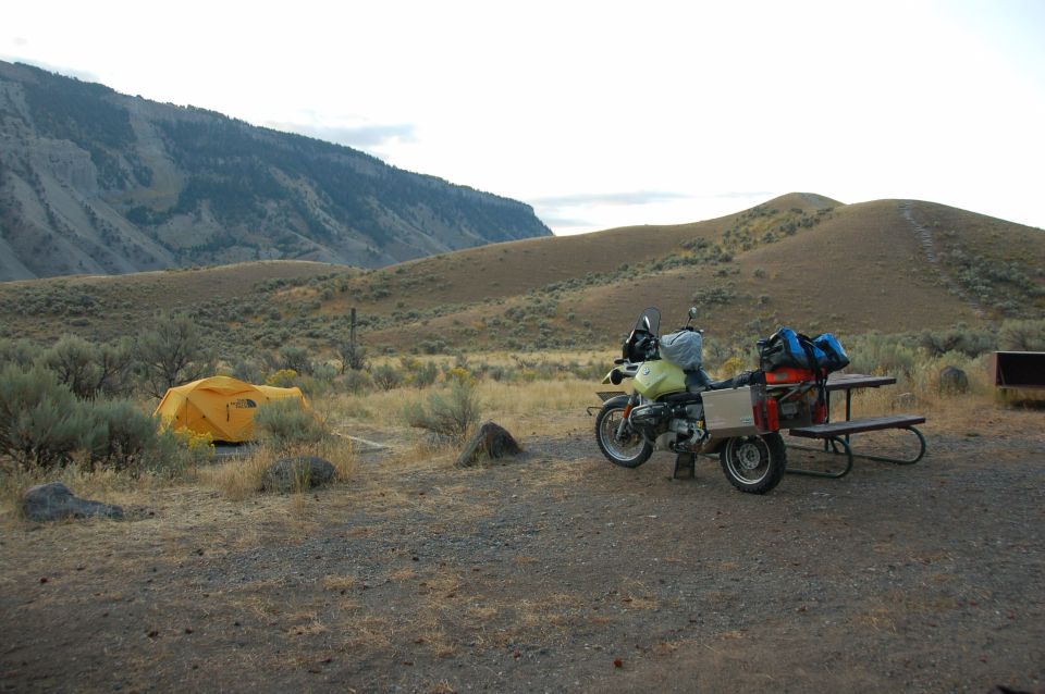 A motorcycle with a trailer is parked at a campsite next to a yellow tent, with hills in the background.