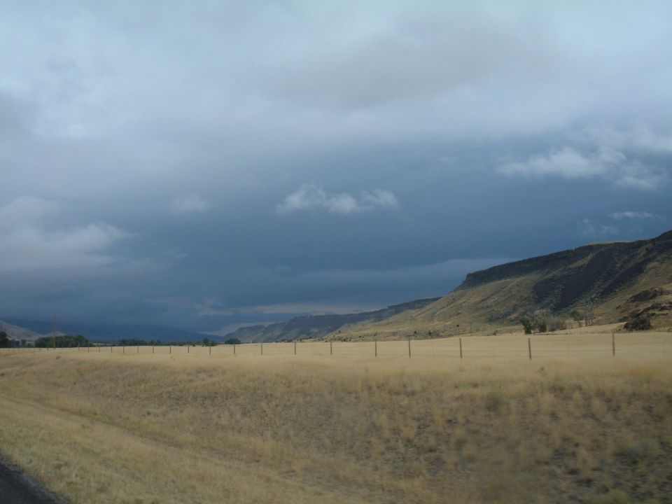 Dry grassy field with a fence and distant hills under a cloudy sky.