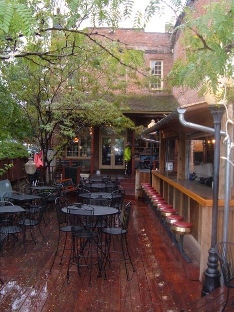 1998 BMW R1100GS ABS - An outdoor patio area with wet wooden decking, tables, and a bar is shown, with a person in a bright yellow jacket visible near an entrance.