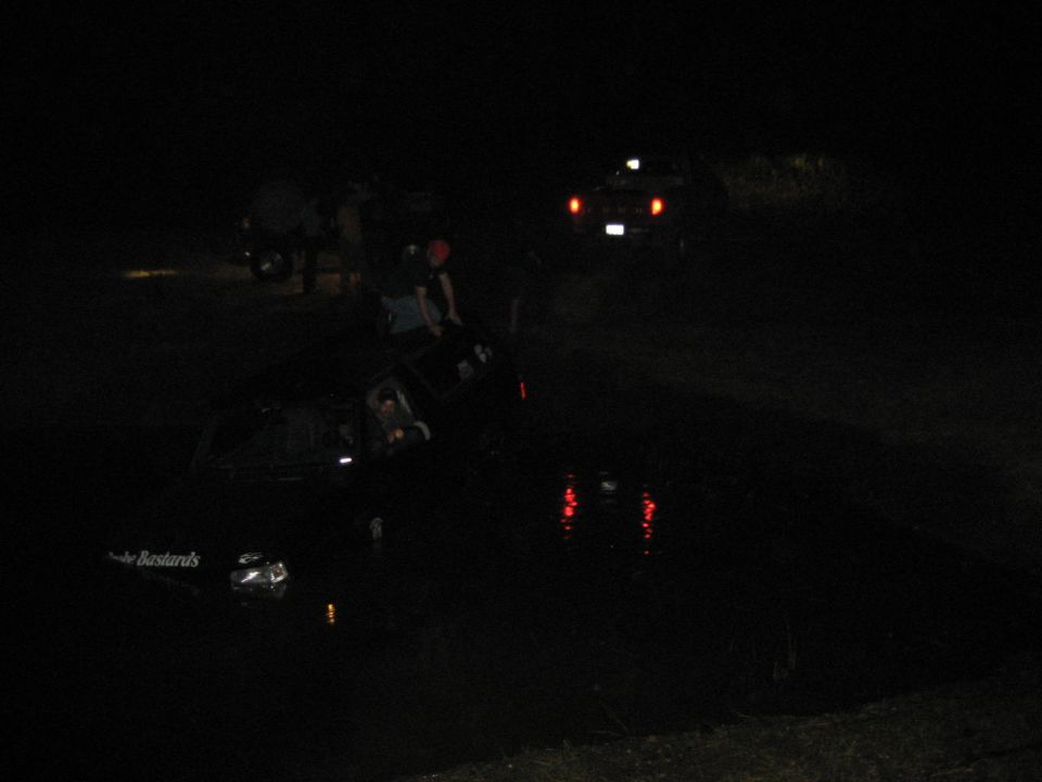 Truck nose-down submerged in a pond at night, tail lights reflecting, onlookers and another truck on the bank